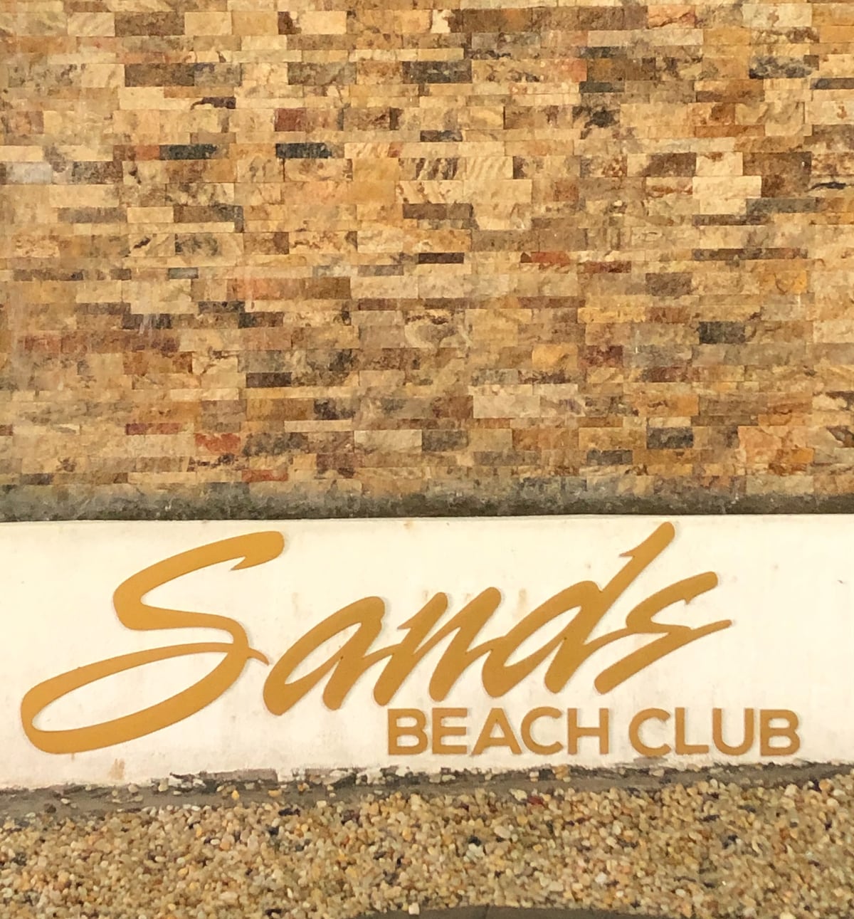 The exterior wall features a textured stone design in warm tones, showcasing the Sands Beach Club sign in elegant gold lettering against a light-colored base. The setting reflects a welcoming atmosphere for guests arriving at the resort.