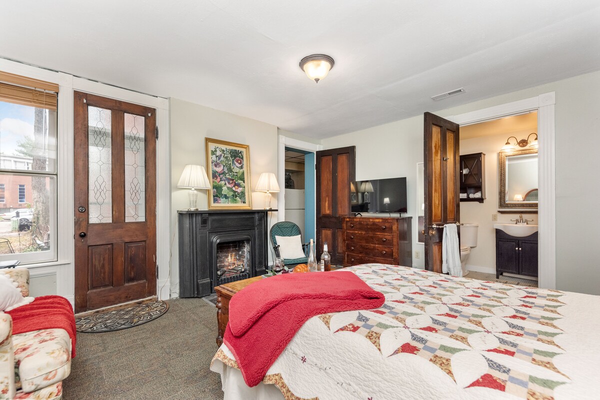 A cozy efficiency apartment is showcased with a colorful quilt draped over a spacious bed. A seating area is provided with a plush chair and decorative couch. The room features two sets of doors leading to the outside and a bathroom visible through the open doorway.