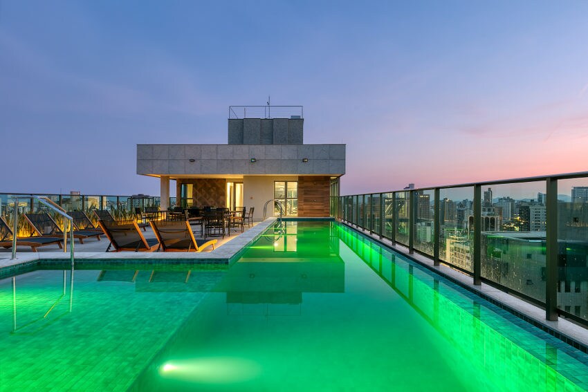 The rooftop pool area features a stunning infinity pool with vibrant green lighting. Comfortable lounge chairs are arranged around the pool, and a communal dining space is visible. The panoramic city views create an inviting atmosphere for relaxation and leisure.