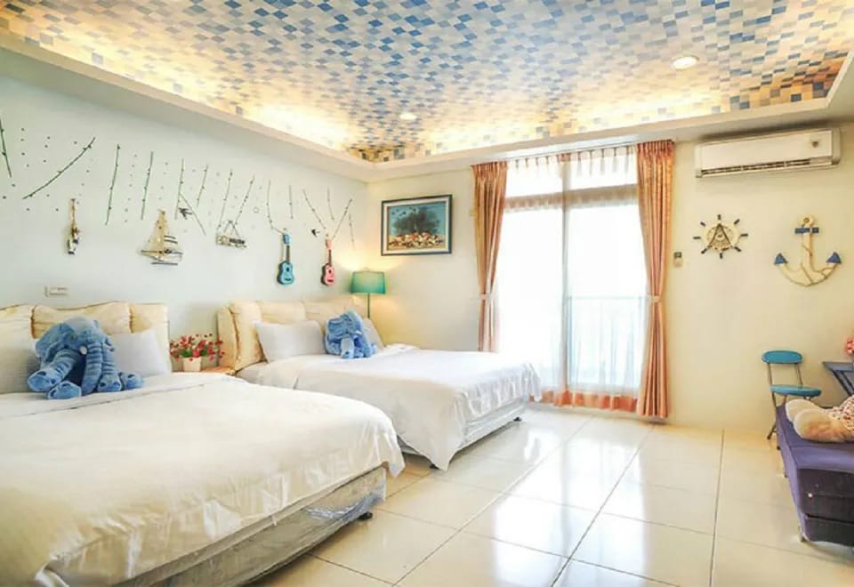 A bright bedroom features two spacious beds adorned with decorative blue and white accents. Wall art includes nautical themes, while large windows with sheer curtains allow natural light to fill the room, complemented by a colorful ceiling design.