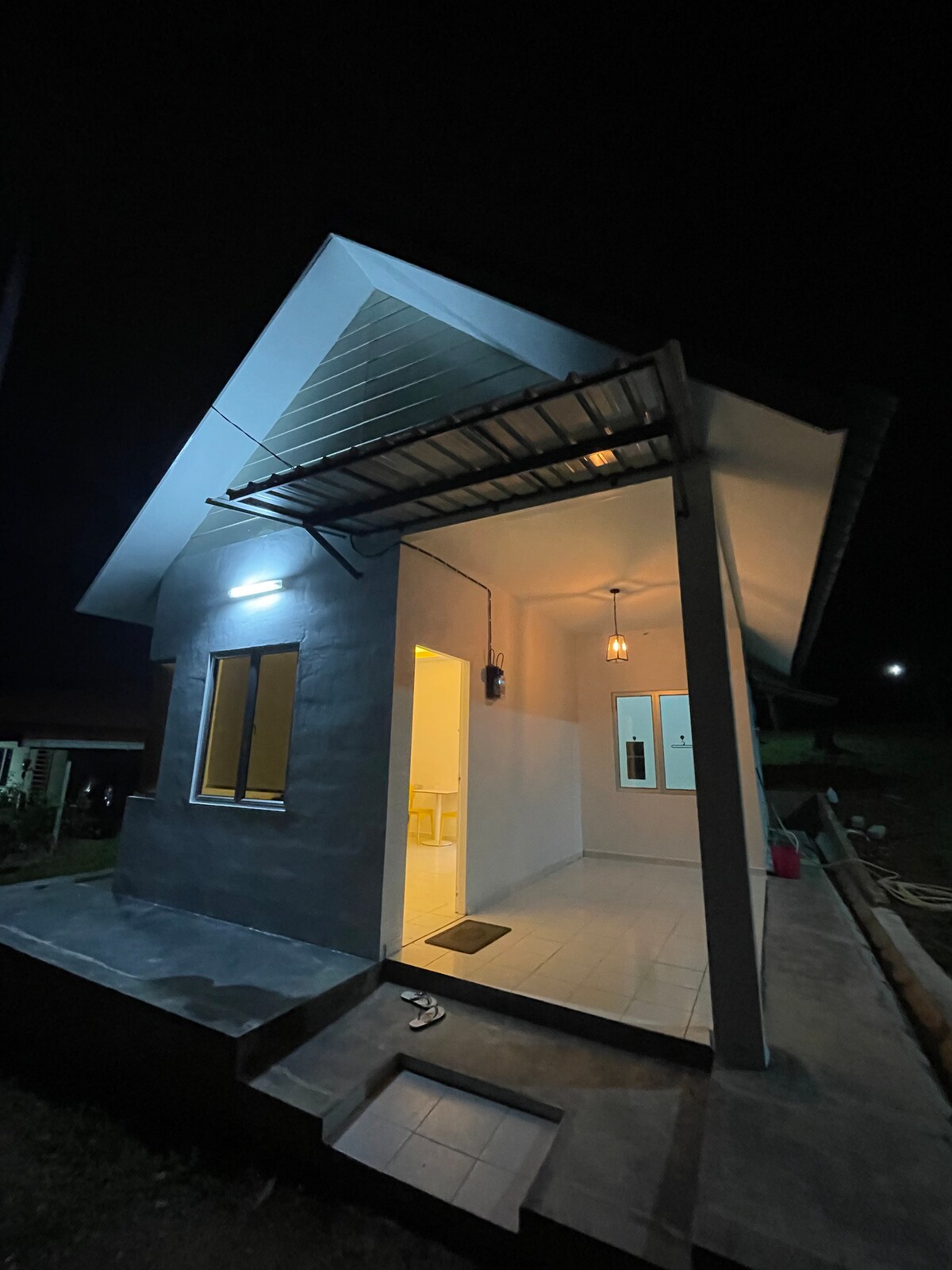 The exterior of the homestay is shown at night, illuminated by warm lighting. A covered entrance leads into the interior, with a simple flight of steps. The structure features a modern design with a sloped roof, complemented by large windows allowing for natural light.