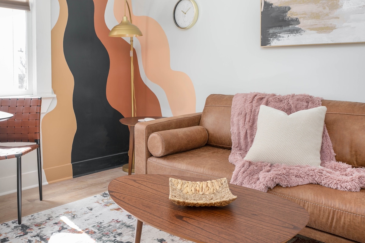 The living area features a contemporary leather sofa complemented by a plush throw pillow and a soft blanket draped over one arm. A round wooden coffee table is positioned in front, with a decorative bowl atop. A wall art piece and a clock are visible, along with a striking painted backdrop.