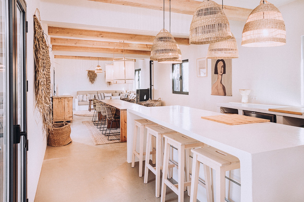 An open-concept living space is shown, featuring a long white island with wooden stools and stylish pendant lights overhead. Natural textures are present through woven elements on the walls. A dining area with a large wooden table and seating is visible in the background.