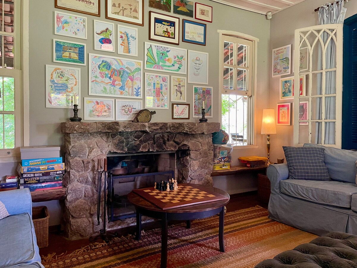 A cozy living area features a stone fireplace framed by a collection of children's artwork adorning the walls. Comfortable, cushioned sofas are positioned around a wooden coffee table, which displays a chessboard. Natural light fills the space through large windows.