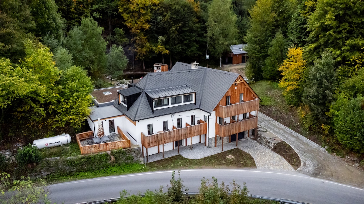 Aerial view of a spacious building with a combination of white and wooden exterior. The structure features multiple balconies and a scenic surrounding of trees in various shades of green and yellow. A winding road leads up to the property, enhancing its natural setting.