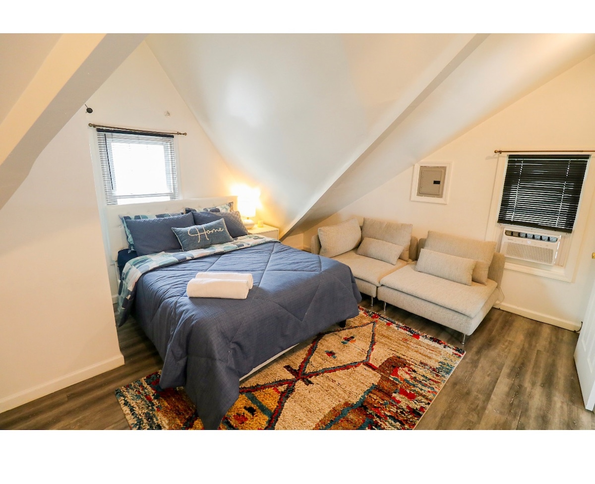 The cozy bedroom features a spacious bed adorned with a textured blue coverlet. A comfortable seating area includes light-colored couches. Natural light fills the room through a window with dark blinds, and a vibrant area rug adds warmth to the wooden floor.