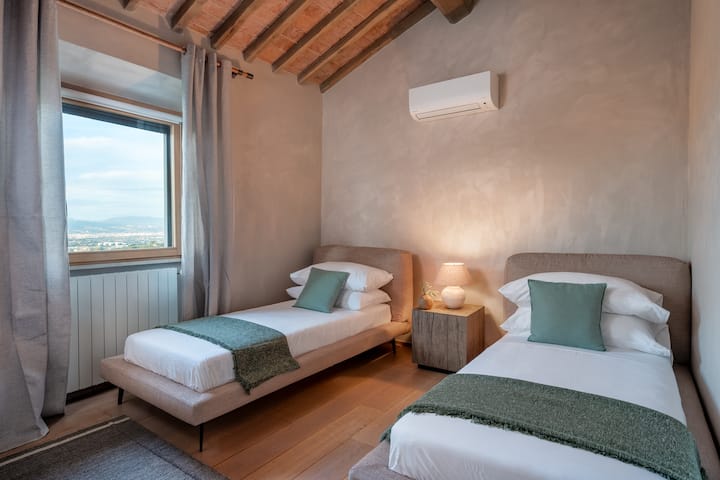 El dormitorio Montalcino cuenta con dos camas individuales, vigas de madera expuestas y vistas serenas de las colinas de la Toscana, que ofrecen un refugio cómodo y elegante en el segundo piso, ideal para niños o huéspedes adicionales.