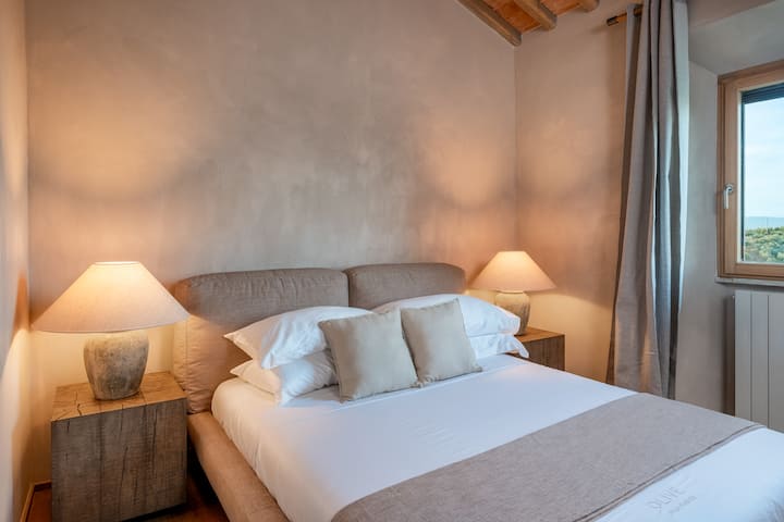 El dormitorio Monticchiello cuenta con una cama tamaño queen, vigas de madera expuestas y vistas relajantes de las colinas de la Toscana, que ofrecen un refugio cálido y tranquilo en el segundo piso de la villa.
