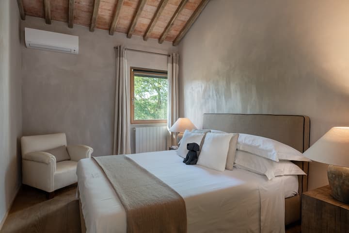 Un dormitorio queen sereno en O:live Villa Firenze, con vigas de madera expuestas, tonos neutros suaves y una ventana que enmarca las vistas panorámicas de las colinas toscanas. Un refugio tranquilo y elegante diseñado para noches de descanso en el campo.