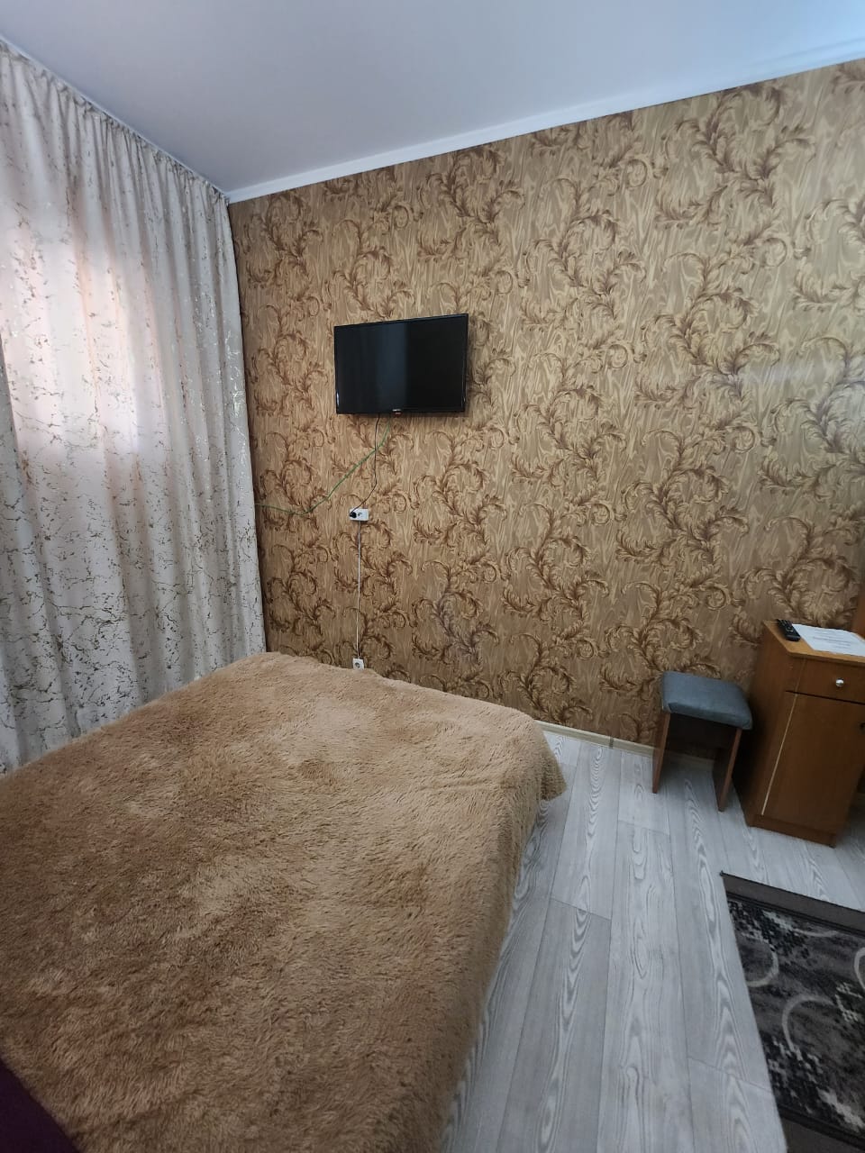 A cozy bedroom features a warm-toned wall with elegant patterns. A spacious bed covered with a soft blanket is positioned near a window dressed with light, flowing curtains. A small desk with a chair is nearby, with a flat-screen TV mounted on the wall.