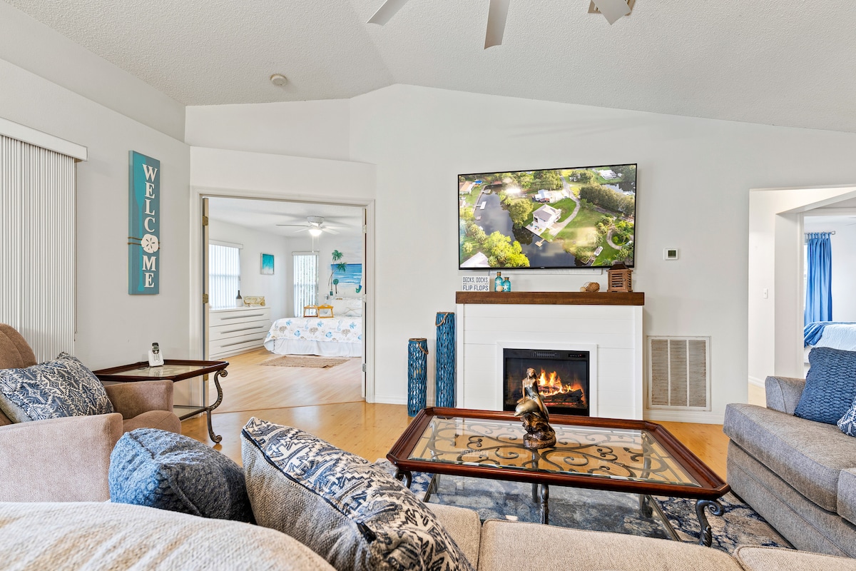 A spacious living area is highlighted by a large flat-screen TV mounted above a white mantle. A comfortable seating arrangement features two sofas adorned with decorative cushions. Natural light is welcomed through sliding glass doors leading to a well-lit bedroom area in the background.