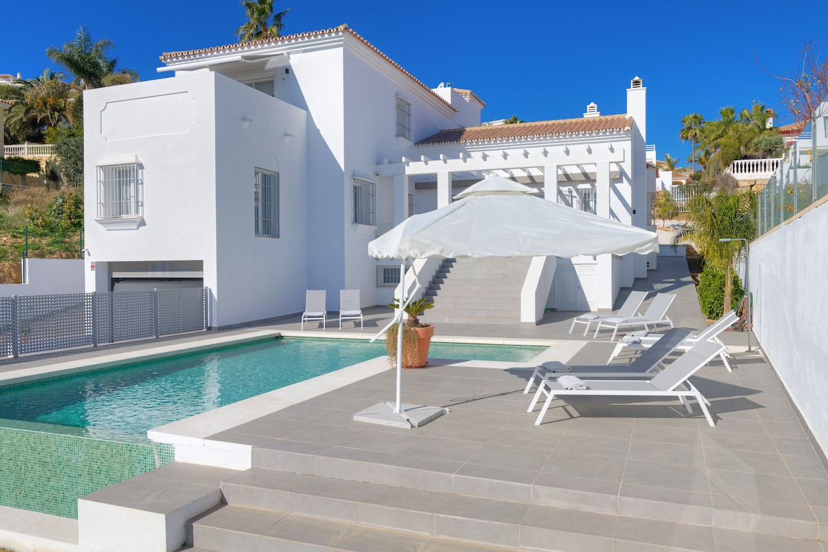 The exterior of the villa is highlighted, showcasing a sleek white facade under a clear blue sky. An infinity pool glimmers beside a spacious terrace, equipped with lounge chairs. A large umbrella offers shade, adding to the inviting outdoor space.