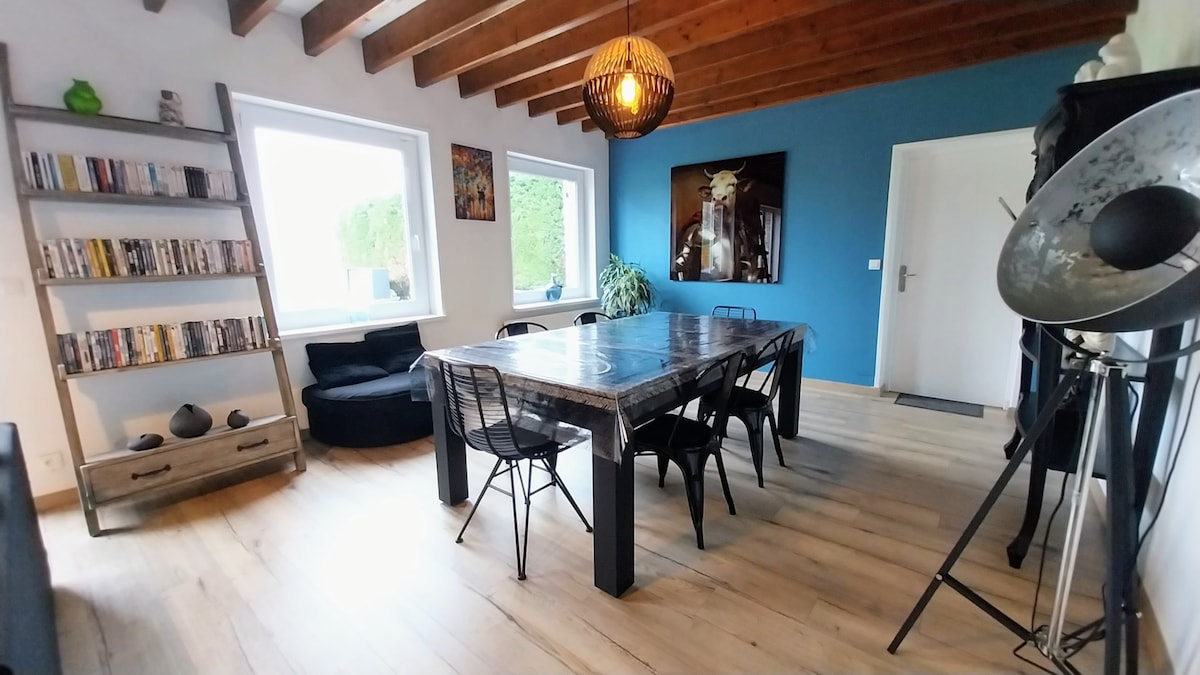 A spacious dining area features a large wooden table surrounded by black chairs. Natural light fills the room through two windows, with a cozy lounge chair placed nearby. A bookshelf holds a collection of DVDs, and artwork adorns the walls.