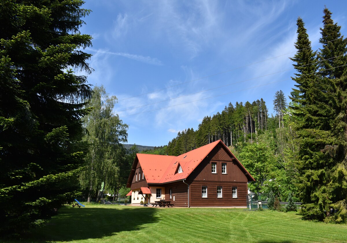 A spacious wooden building with a striking red roof is surrounded by lush greenery and tall trees. The structure features multiple windows and a large outdoor area with well-maintained grass. Blue skies and wispy clouds are visible above, enhancing the serene environment.