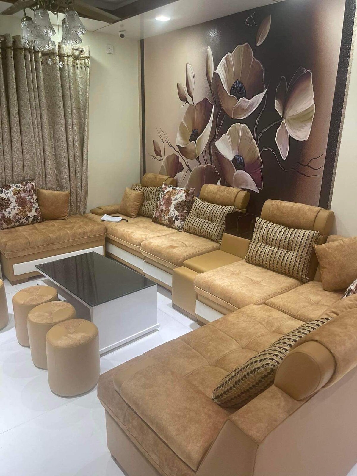 A spacious living area features a large sectional sofa adorned with decorative pillows, complemented by elegant stools. A chic coffee table occupies the center, while a floral wall mural provides a tasteful backdrop. Soft curtains frame the window, enhancing the room's cozy feel.
