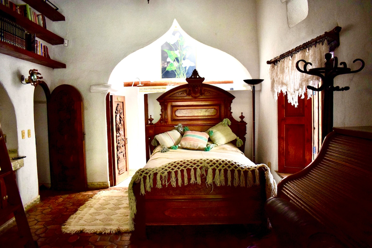 A spacious bedroom is highlighted by an ornate wooden bed adorned with pillows and a knitted throw. Natural light filters through an arched window above, illuminating the room's adobe walls. An antique chair and a decorative coat rack add character to the cozy space.