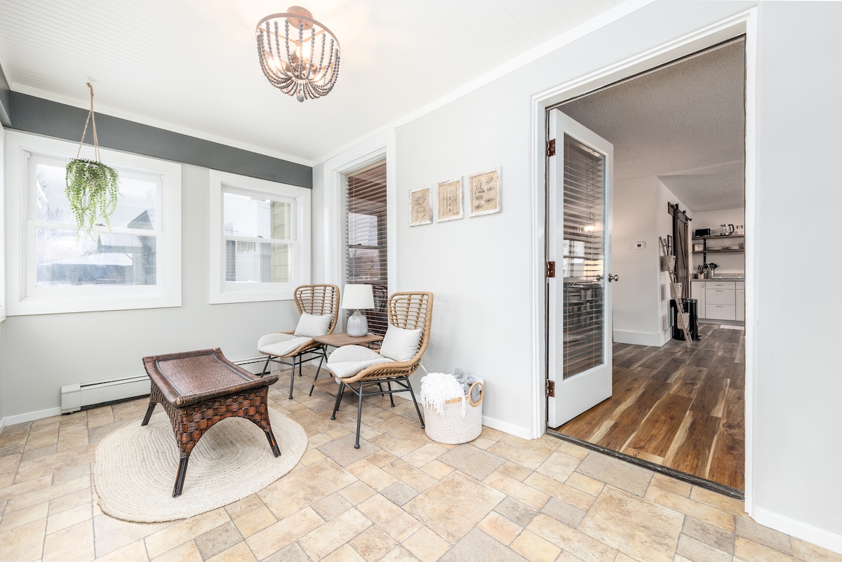A bright sunroom showcases two rattan armchairs arranged around a small woven coffee table. A round area rug lies on the tile floor, and large windows allow natural light to fill the space. Access to the adjoining living area is visible through an open door.