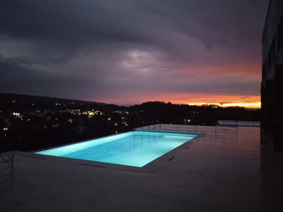 A serene pool area reflects the soft glow of evening light, with a backdrop of a colorful sunset illuminating the sky. The expansive pool is surrounded by a clean, smooth surface, enhancing the view of the surrounding hills and distant city lights.