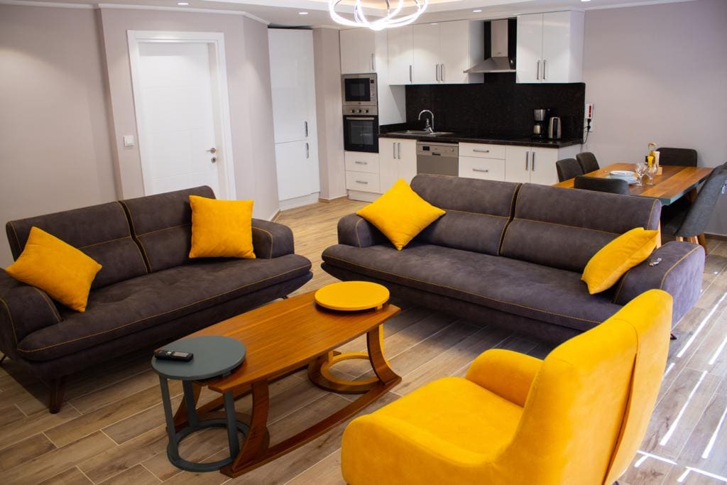 A modern living area features two dark gray sofas, adorned with yellow cushions, positioned around a circular wooden coffee table. A light-colored dining table is visible in the background, complemented by stylish dining chairs. Ample natural light illuminates the space from a nearby window.