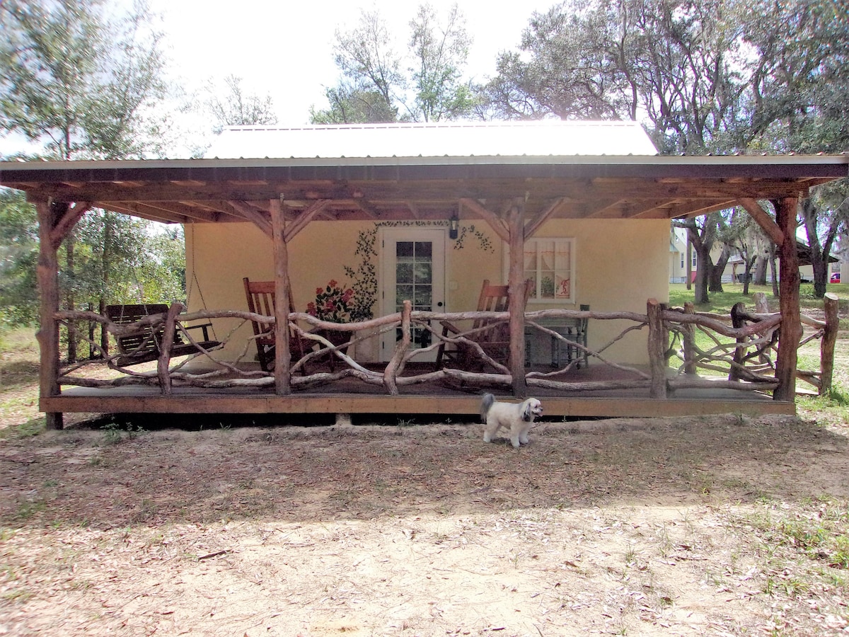 A cottage exterior is presented, featuring a spacious porch supported by rustic wooden posts. Rocking chairs are positioned along the railing, and a small dog is seen on the ground. The charming façade includes soft landscaping and surrounding trees.