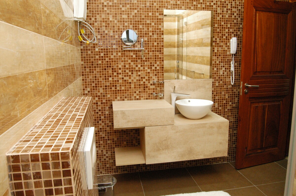 The bathroom features a tiled wall adorned with mosaic patterns, complementing a modern, minimalist vanity with a round sink. A large mirror is mounted above the countertop, and a warm wooden door adds a contrasting element to the serene decor.