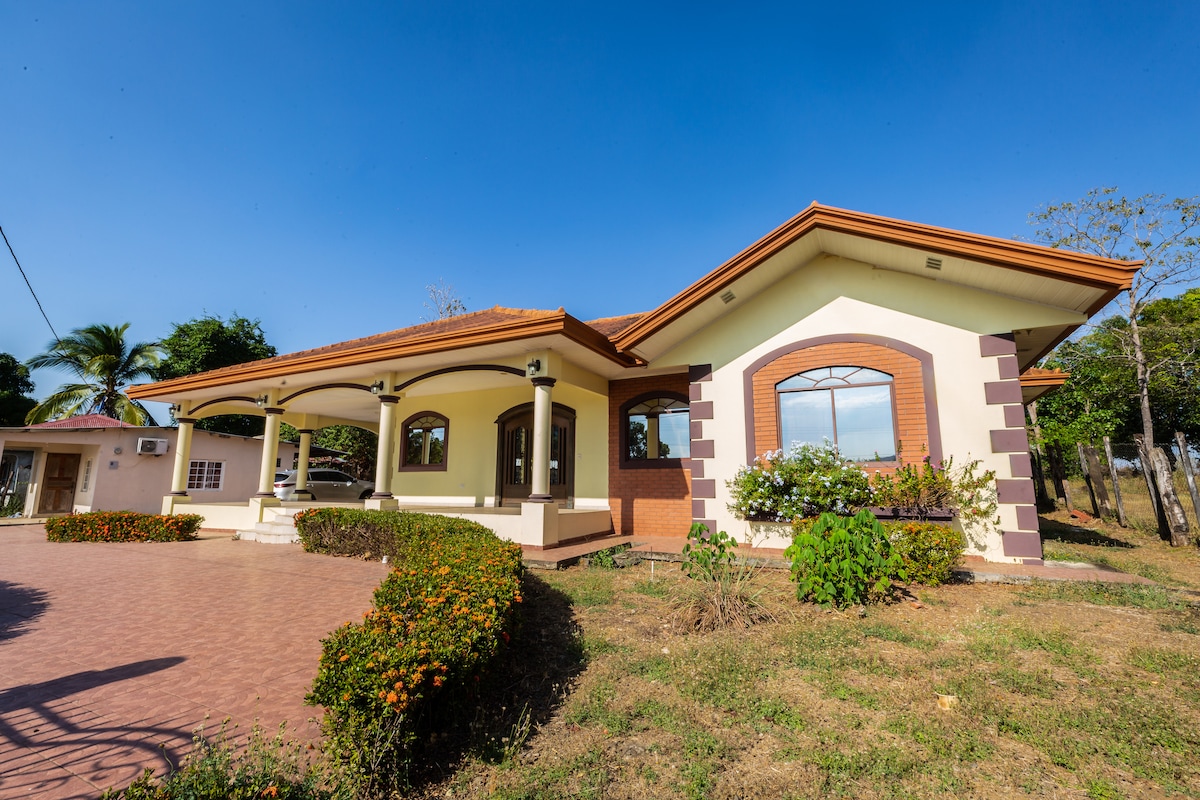 A charming house with a tiled roof and arched windows is set against a clear blue sky. Lush greenery surrounds the exterior, while the driveway is lined with flowering bushes. The welcoming entryway features wide steps leading up to the covered porch.