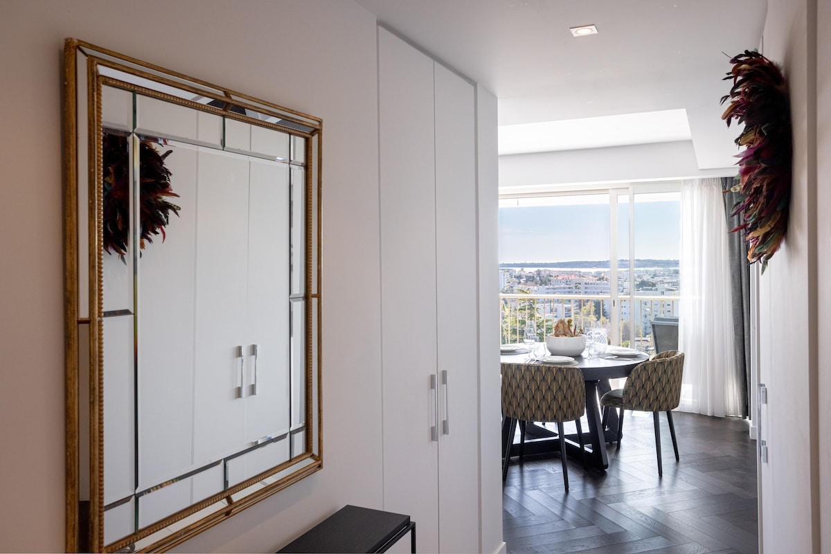 A hallway features a large decorative mirror with a gold frame reflecting natural light. White storage cabinets line one side, complementing the sleek black furniture. At the end, a dining area with a round table and patterned chairs offers a view through expansive glass doors.