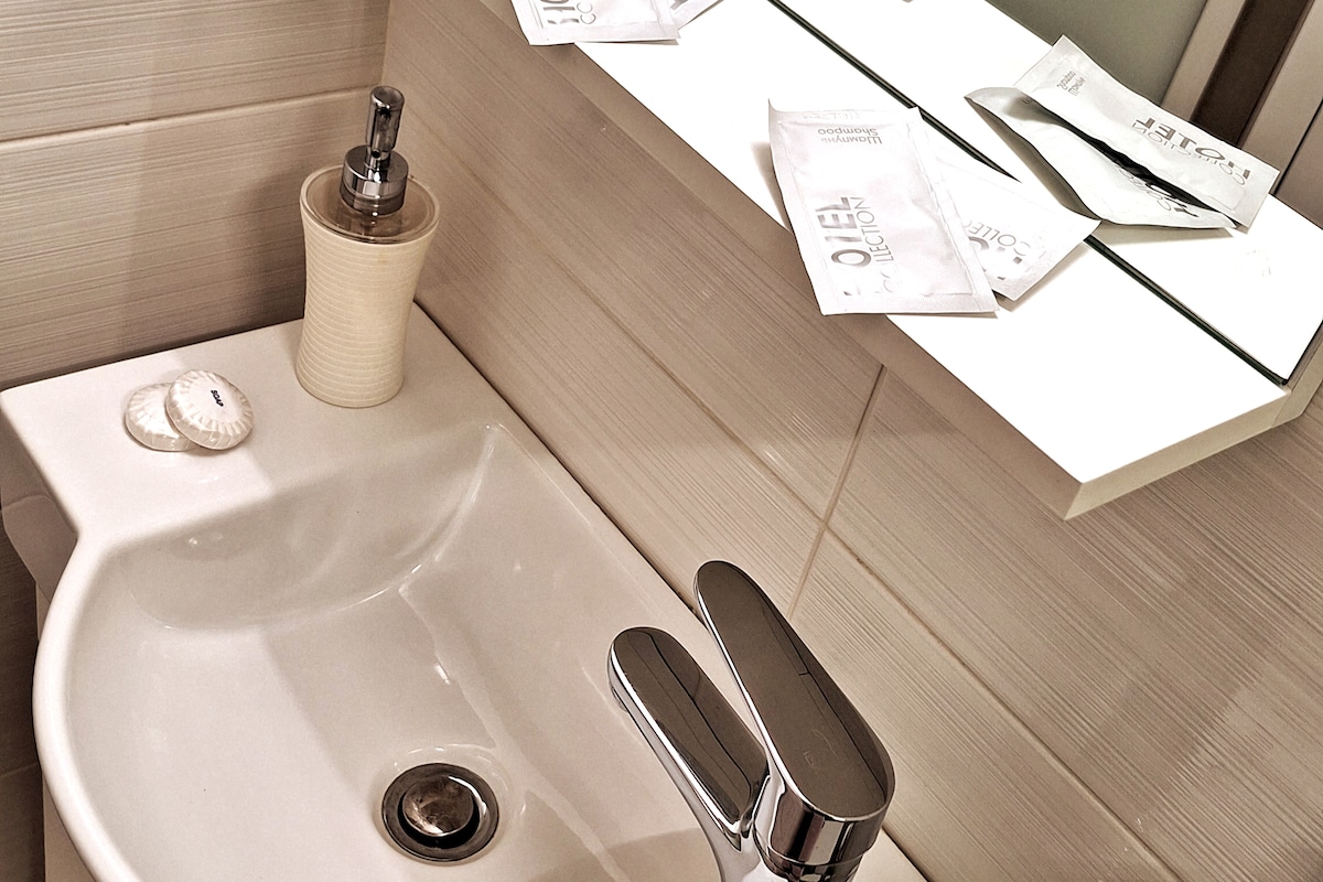 A modern bathroom sink is shown, featuring a sleek faucet and a ceramic soap dispenser. The countertop is arranged with neatly placed individual toiletries and a reflective mirror above. Soft lighting highlights the clean lines and neutral color palette of the space.