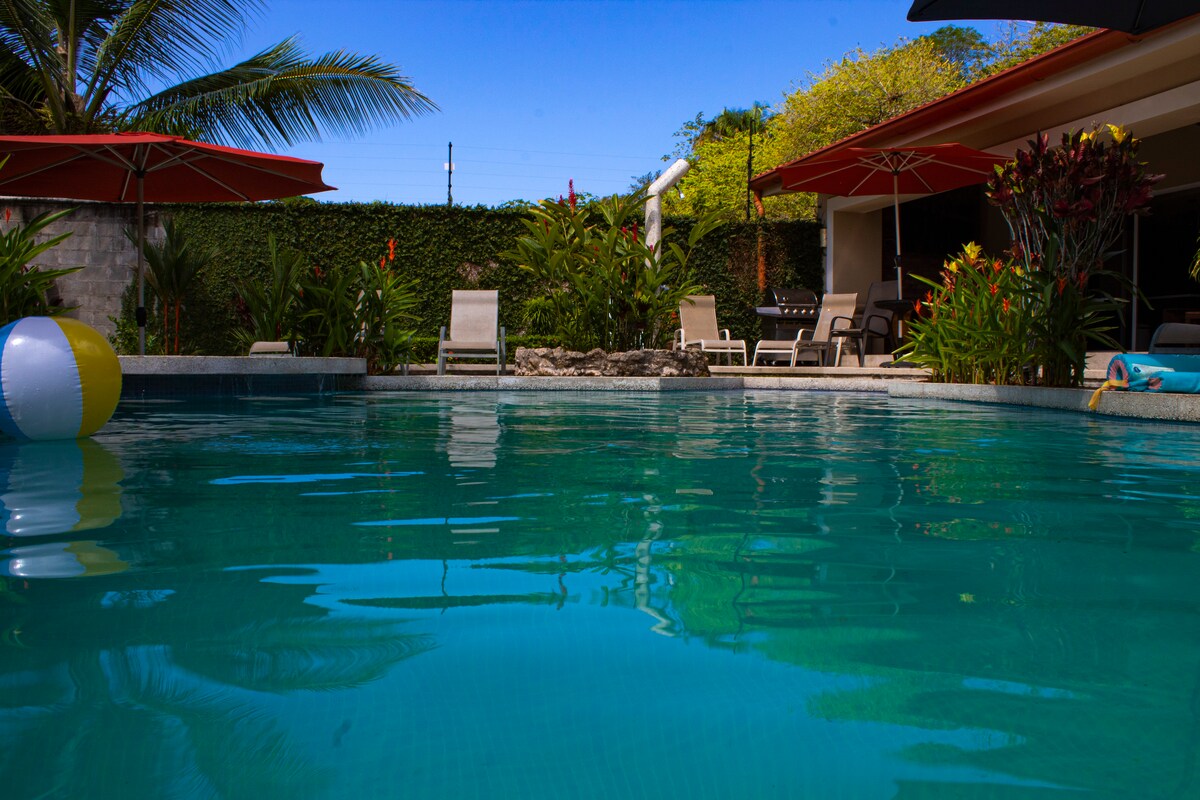 A private pool is surrounded by lush greenery, with vibrant tropical plants and a few lounge chairs positioned along the water's edge. Colorful umbrellas provide shade, and a beach ball floats gently on the clear water under a bright blue sky.