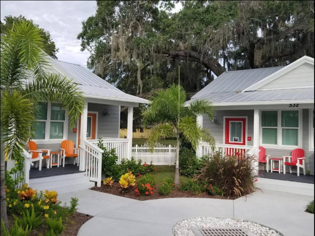 Two charming cottages are featured, each with a welcoming porch surrounded by tropical landscaping. Brightly colored chairs are placed on the porches, while lush greenery and vibrant flowers enhance the inviting outdoor space.