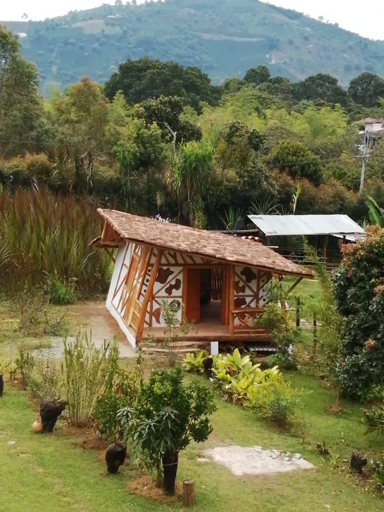 A charming cabin with a sloped roof and decorative wooden accents sits amidst lush greenery. The structure is surrounded by a well-maintained garden featuring various plants and trees. Rolling hills are visible in the background, enhancing the natural setting.