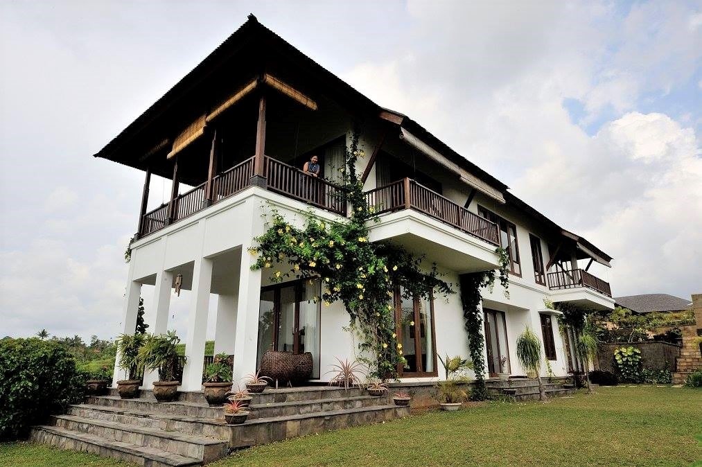 A two-story villa is surrounded by lush greenery and features a spacious outdoor area. Large windows and multiple balconies enhance the open feel, while potted plants add natural charm to the entrance. The structure's roof is adorned with traditional Balinese design elements.