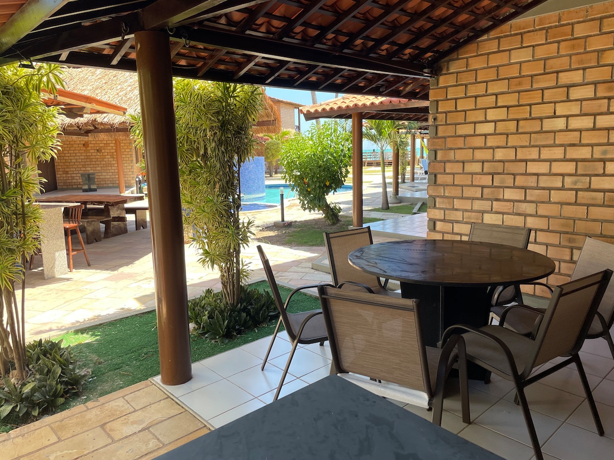An outdoor seating area features a round table surrounded by several cushioned chairs. Lush greenery frames the space, with a view of a pool in the distance, complemented by a covered patio roof and tiled flooring.