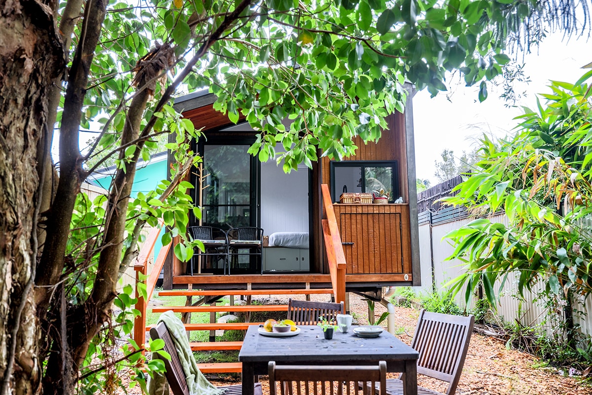 A wooden deck features a set of outdoor dining furniture shaded by lush foliage. The Tiny Home's entrance is framed by greenery, showcasing large glass doors that invite natural light into the interior. The surrounding area includes vibrant plants, enhancing the tranquil setting.