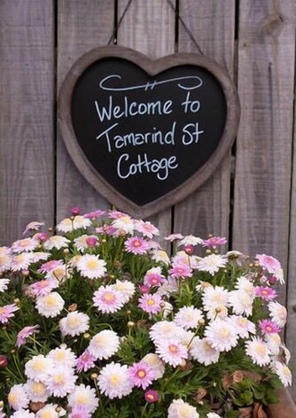A heart-shaped chalkboard sign hangs on a wooden fence, featuring the welcoming message 'Welcome to Tamarind St Cottage.' Below, a vibrant arrangement of pink and white flowers adds a touch of color to the inviting entrance.