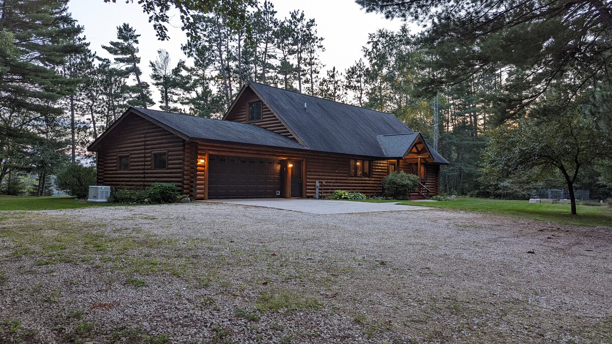 The exterior of a spacious log house is set among tall trees, with a gravel driveway leading up to it. Soft outdoor lighting illuminates the entrance, which features a covered porch. The surrounding lush landscape provides a serene atmosphere.
