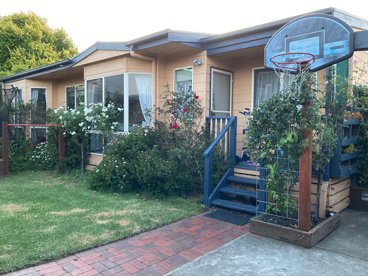 The exterior of the guest house features a landscaped area with flowering bushes and a manicured lawn. A paved pathway leads to a blue wooden entrance, complemented by a basketball hoop mounted by the porch. Large windows invite natural light into the living space.