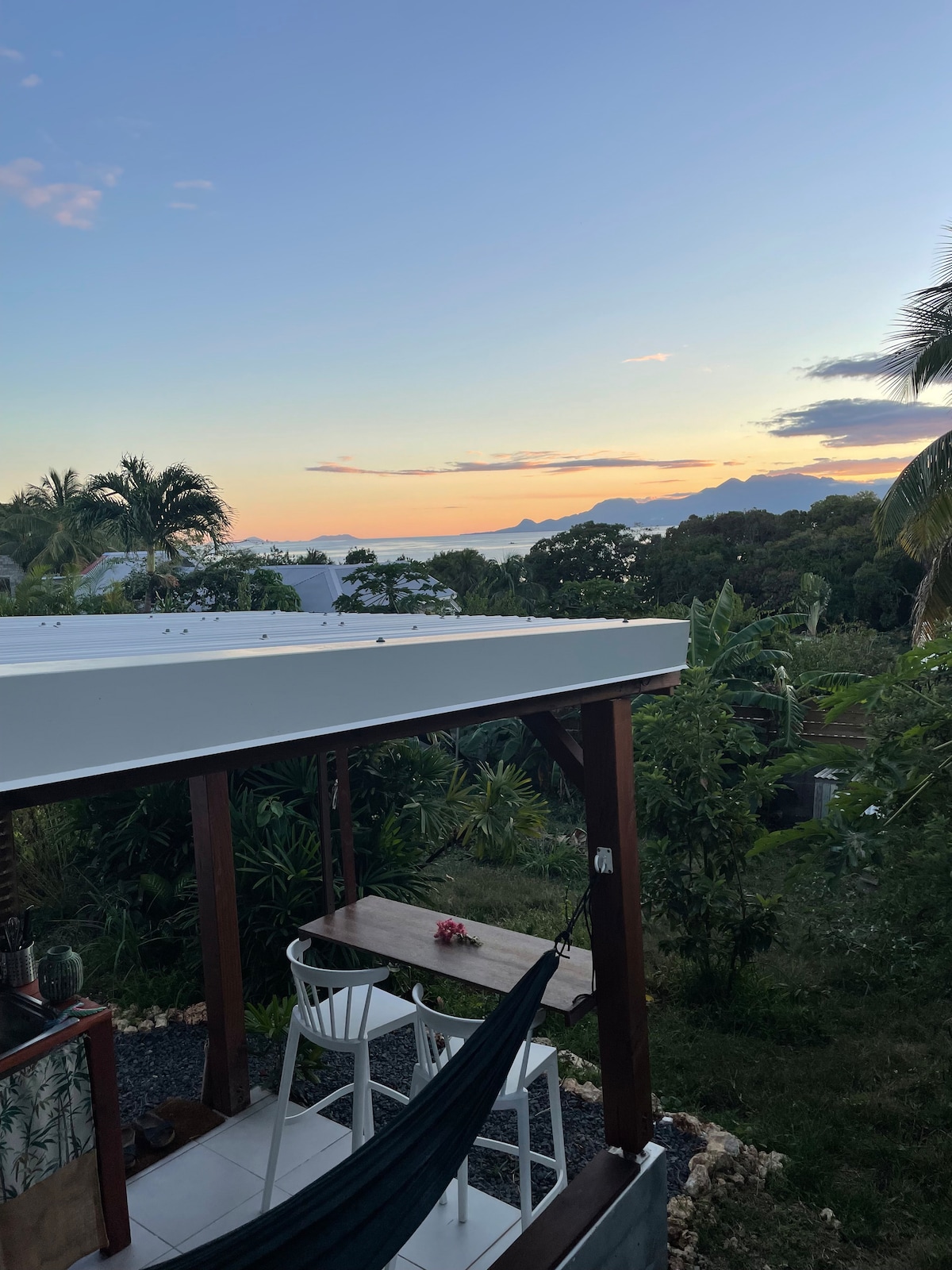 A terrace area is visible, featuring a high table with two chairs and a hammock suspended nearby. Lush greenery surrounds the space, with a view of the ocean and mountains in the distance. The sky is illuminated by soft colors as the sun sets.