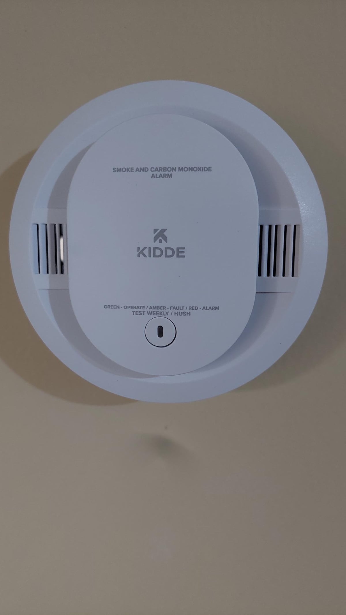 A smoke and carbon monoxide alarm is mounted on a light-colored wall. The round device features a central button and ventilation slits on either side, indicating its purpose for safety within the space.