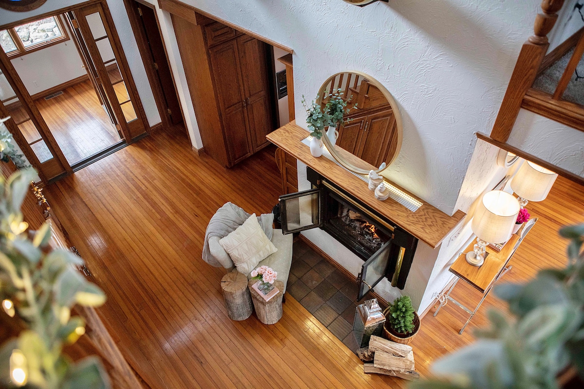 An inviting living area is showcased with a wood-burning fireplace surrounded by natural wood accents. A comfortable seating option is paired with rustic side tables. Large windows allow for ample light, highlighting the rich wooden flooring and open layout of the space.