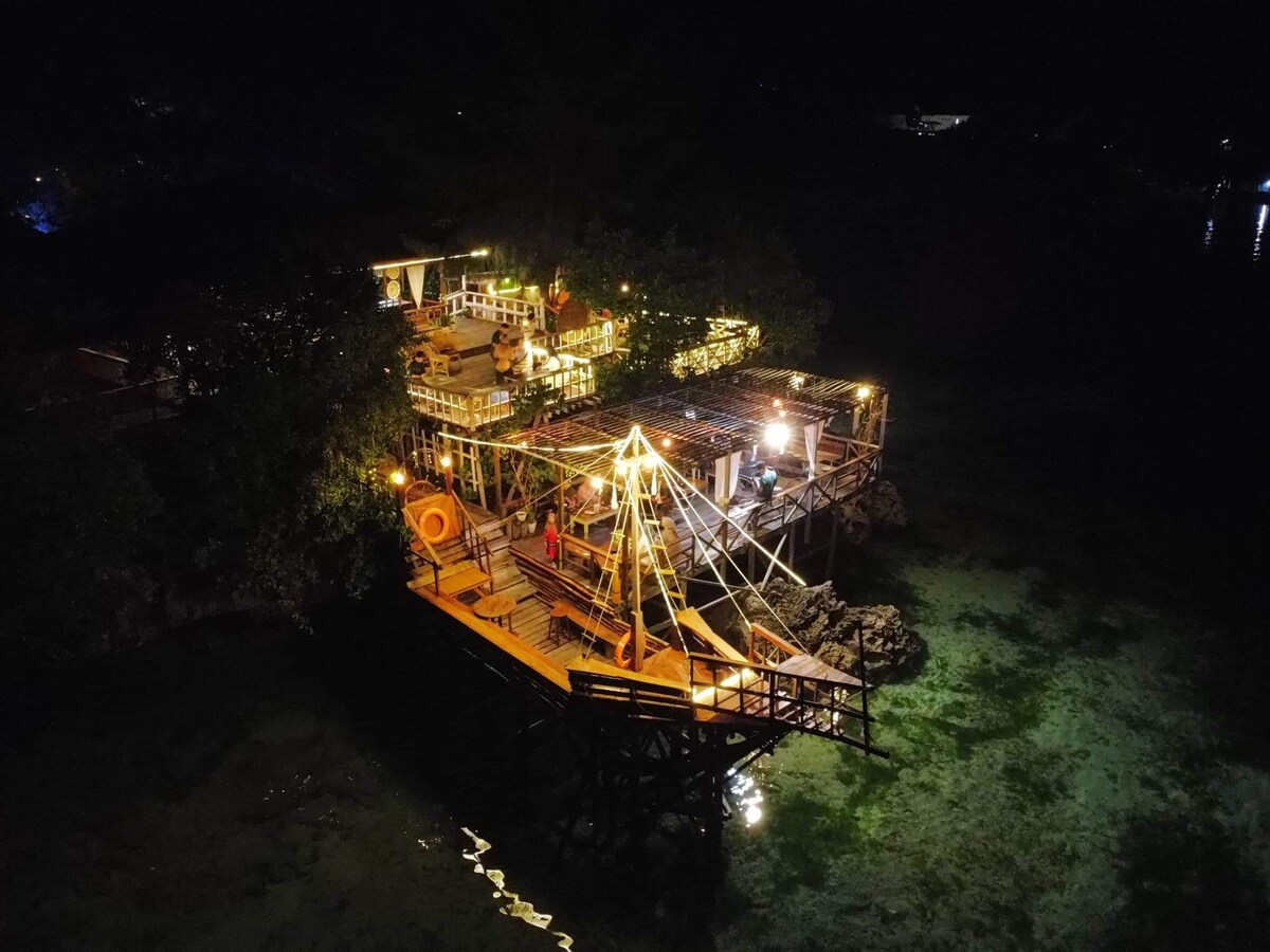 A wooden structure is illuminated at night, showcasing an outdoor dining area with string lights overhead. The building extends over water, surrounded by gentle waves. The natural landscape is visible in the background, enhancing the serene ambiance of the setting.
