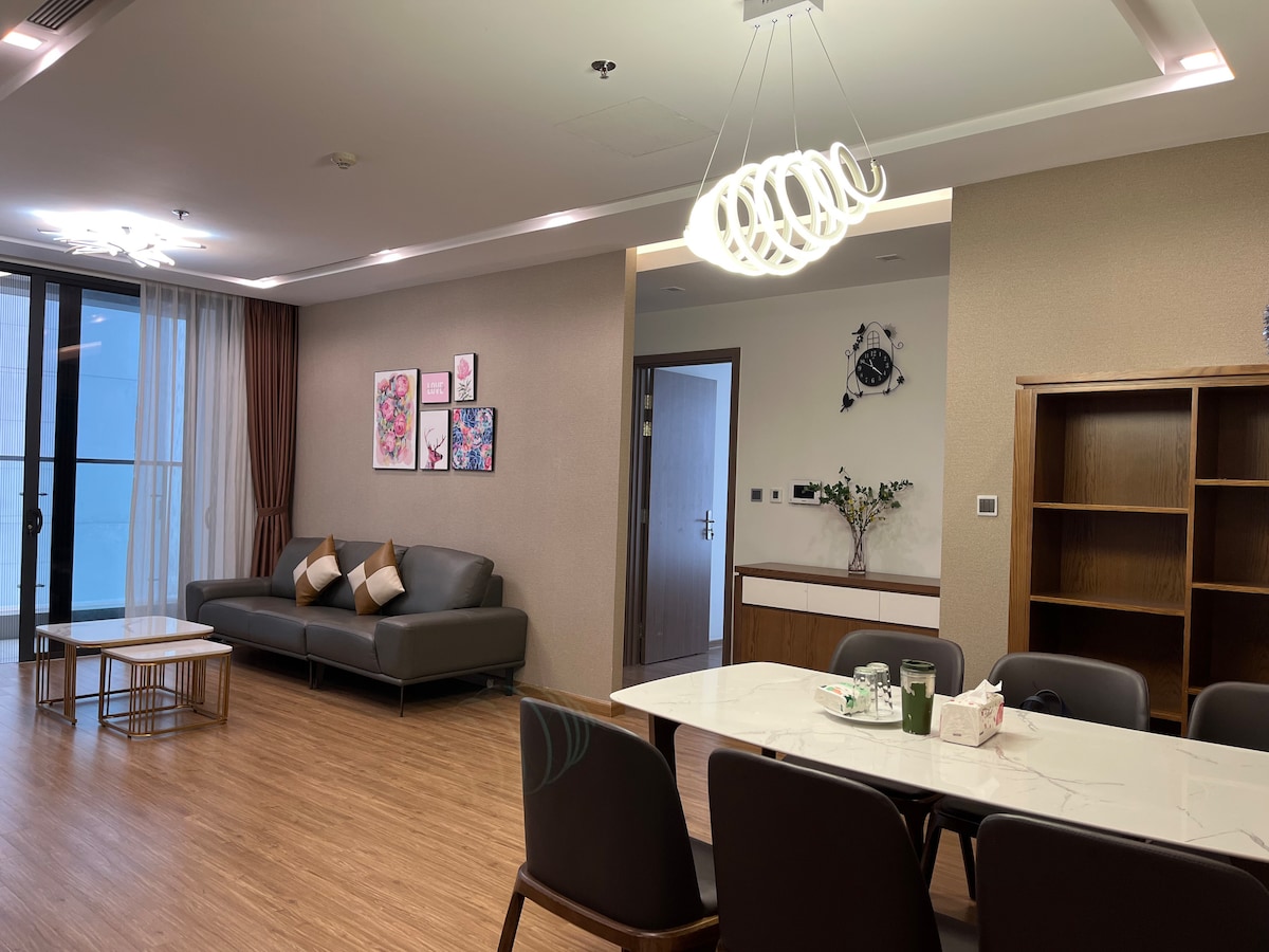 A spacious living area features a dark sofa with patterned cushions, a light-colored coffee table, and a dining table surrounded by chairs. The room is illuminated by a modern ceiling light, with artworks displayed on the walls and sliding glass doors providing natural light.