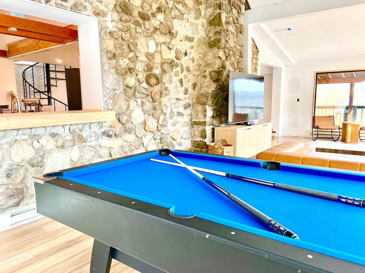 A vibrant blue pool table is highlighted in the foreground, showcasing two cue sticks positioned across its surface. A stone accent wall lines the background, while a large television and comfortable seating area are visible in the open space beyond.