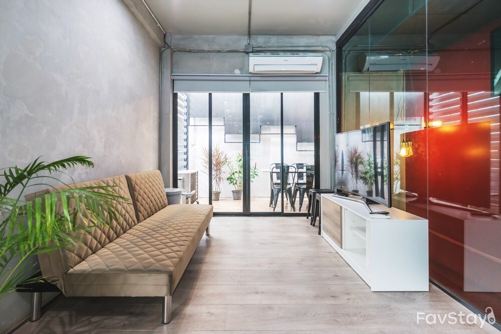 A modern living area features a stylish sofa with diamond-patterned upholstery, alongside a sleek white entertainment unit. Large glass doors open to a patio area, while indoor plants add a touch of greenery to the neutral-toned space.
