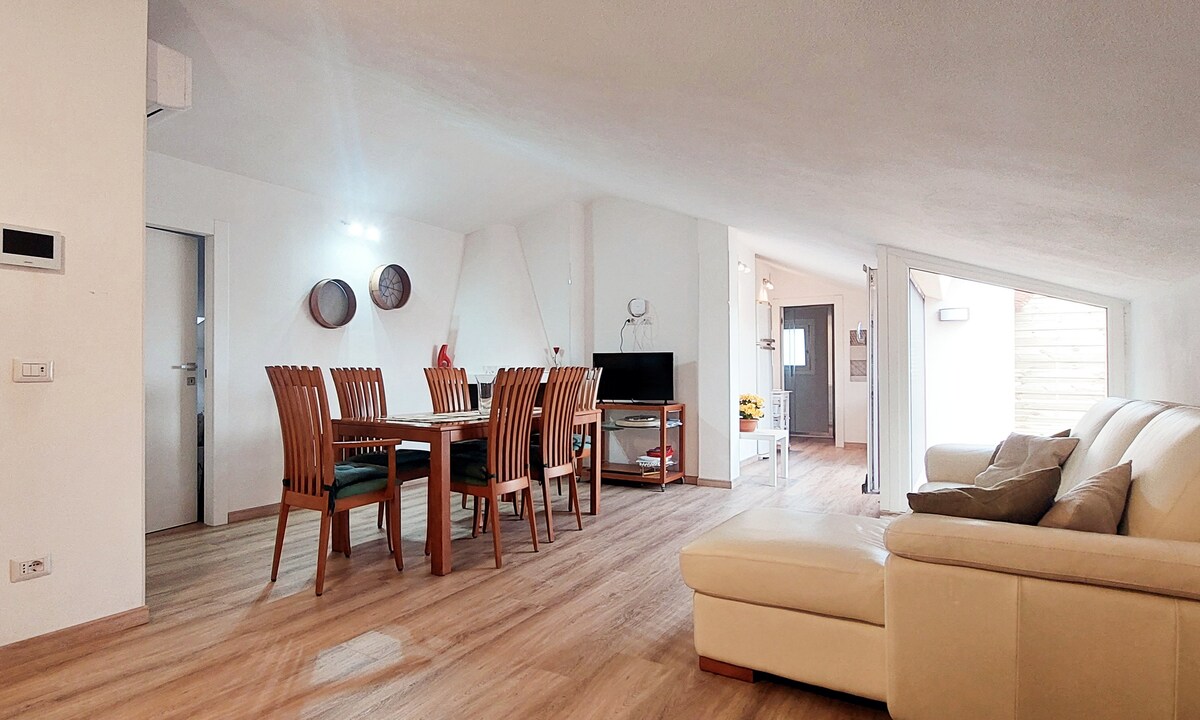 A spacious living area features a comfortable cream-colored sofa and a wooden dining table with multiple chairs. A television is positioned nearby. Natural light floods the space through large windows, creating a bright and inviting atmosphere.