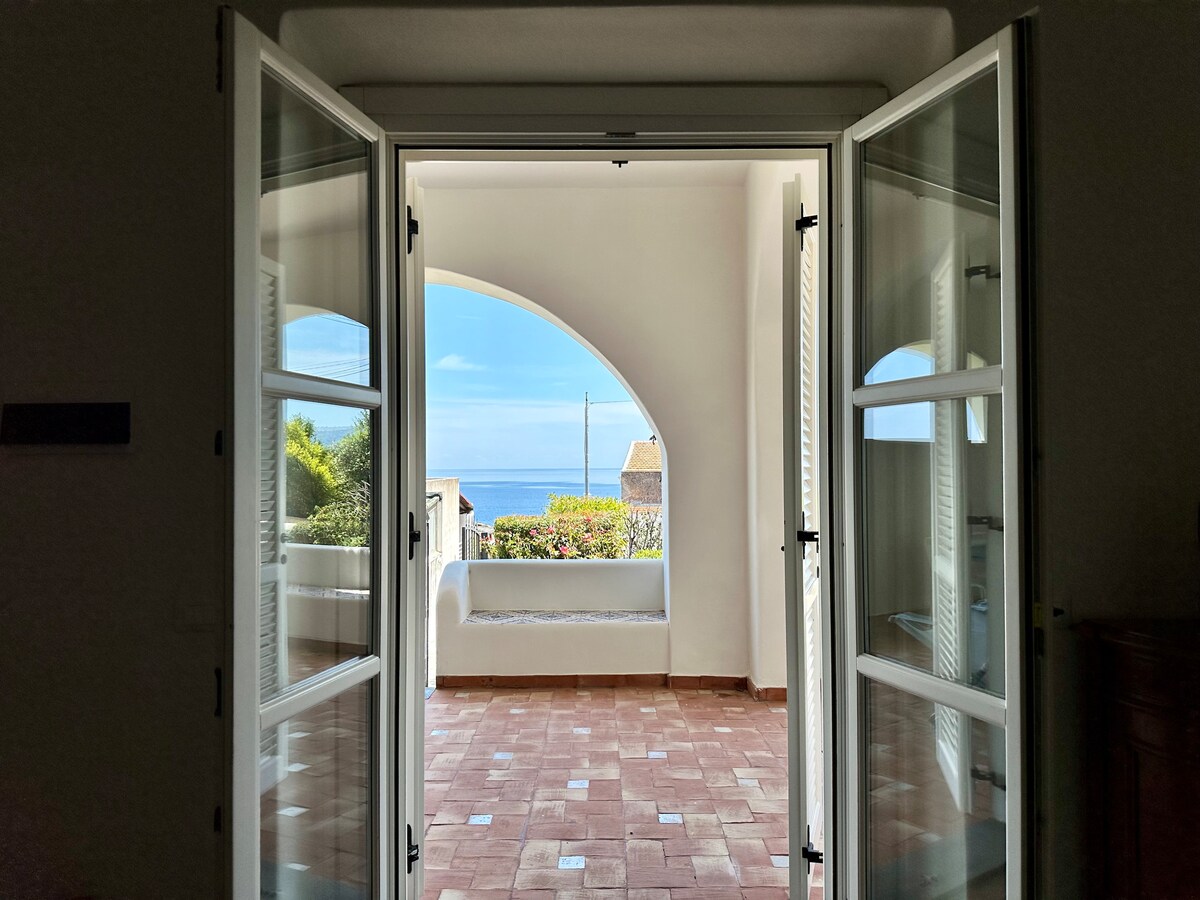 An arched doorway with double glass doors opens to a spacious terrace, offering a partial view of the sea. The floor is tiled in warm terracotta, and sunlit spaces are framed by white walls, creating a bright and airy atmosphere.