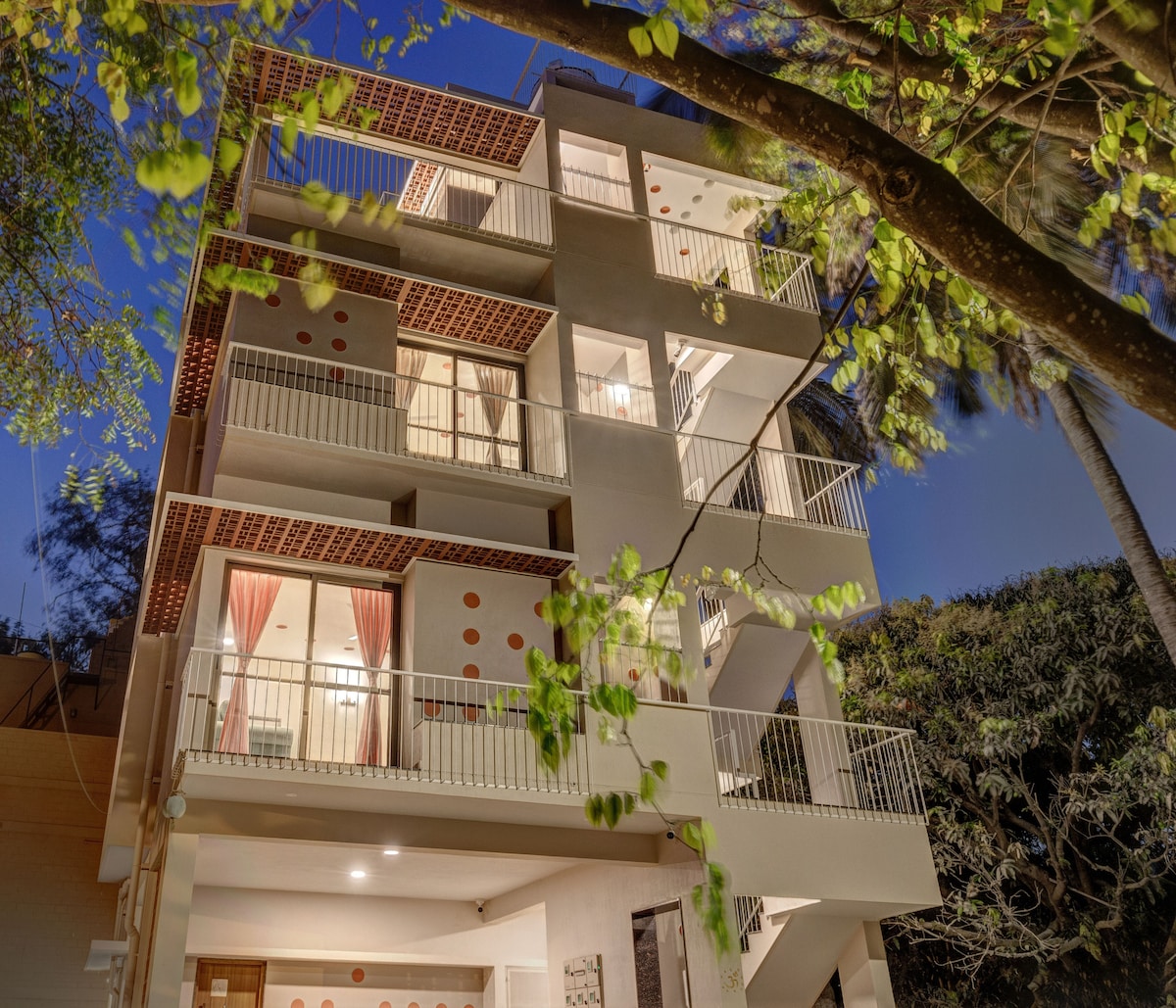 The building features a modern design with multiple balconies, allowing for ample natural light. Lush greenery surrounds the structure, enhancing the sense of privacy. The warm glow from the interior adds a welcoming touch to the evening atmosphere.