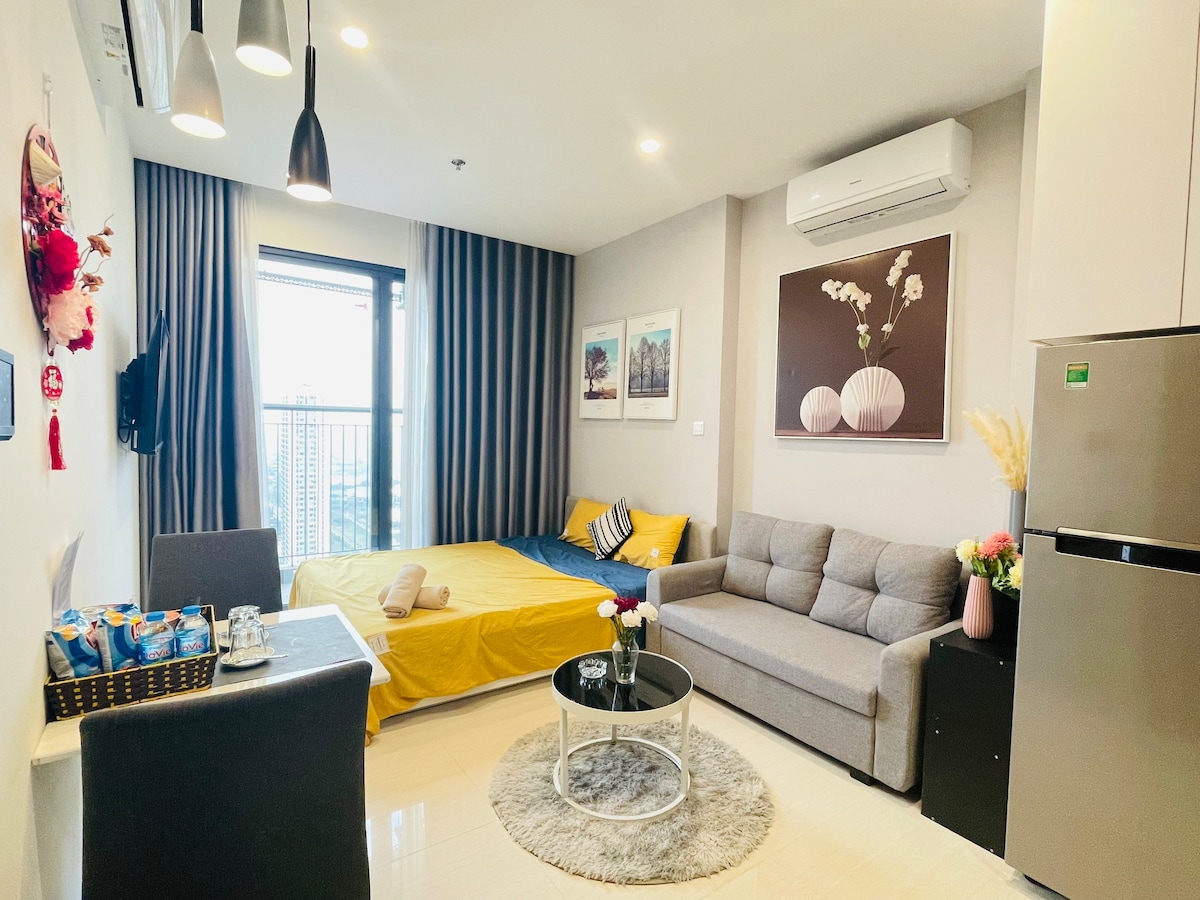 A modern room is displayed, featuring a cozy seating area with a gray sofa and a coffee table. A spacious bed with yellow bedding is visible next to large windows framed by soft curtains, while decorative art adorns the walls. A kitchenette is also present.