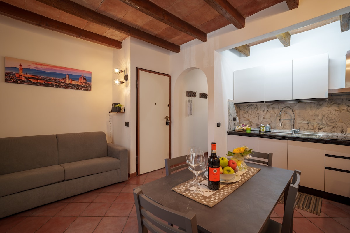 A combined kitchen and living area is highlighted by a grey sofa and a dining table set for four. The kitchen features modern cabinetry, with a stone-like backsplash creating texture. Natural light illuminates the space, complemented by rustic wooden beams overhead.