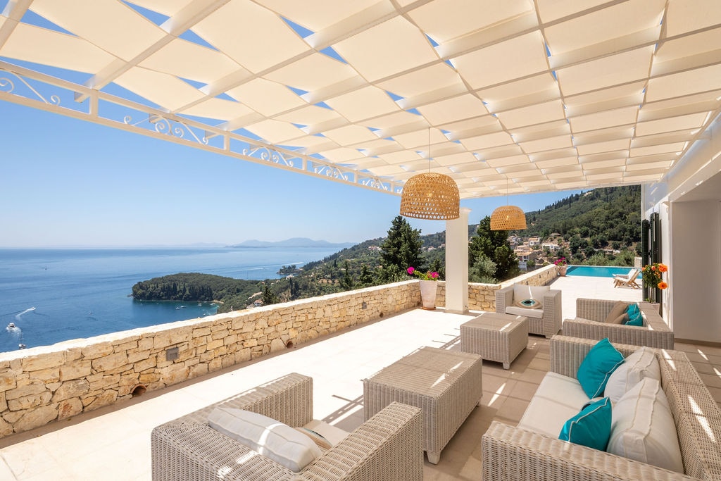 A spacious outdoor terrace features woven furniture arranged for relaxation, complemented by decorative lighting fixtures. The area is shaded by a large canopy, with views of the serene coastline and ocean in the background. Vibrant flowers are visible in the flowerbeds.
